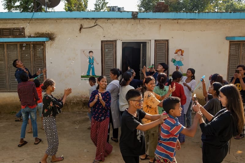 Lots of kids wearing different coloured shirts are playing outside, in front of a white building with images painted on the front.