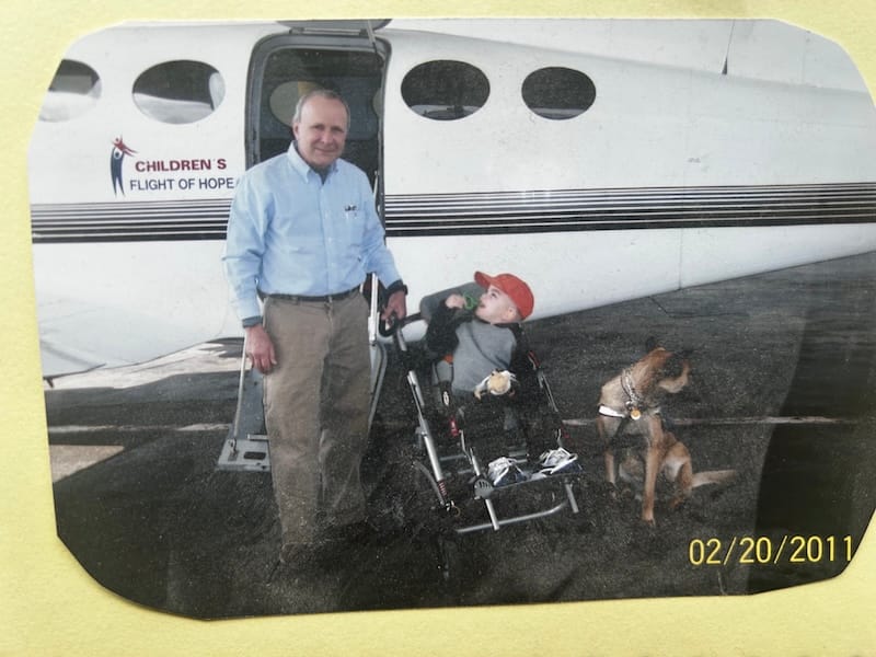 A man stands outside the door of a plane labelled 'Children's Flight of Hope'. Next to him is a young child wearing a red cap, sitting in a pram/wheelchair device. There is also a dog next to the child.